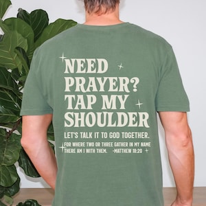 May include: Olive green t-shirt with the text "NEED PRAYER? TAP MY SHOULDER" in a cream-colored font. Below the text is the phrase "LET'S TALK IT TO GOD TOGETHER." and a Bible verse. The shirt is a short-sleeved, casual style.