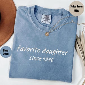 May include: Blue jean colored t-shirt with the words "favorite daughter since 1996" in white script. A gold necklace with a pendant rests on the shirt. The shirt has a Comfort Colors tag.