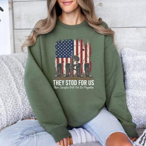 May include: An olive green sweatshirt featuring a distressed American flag design with military boots, a helmet, and a rifle. The text "THEY STOD FOR US" and "Their Sacrifice Will Not Be Forgotten" is printed below the graphic.