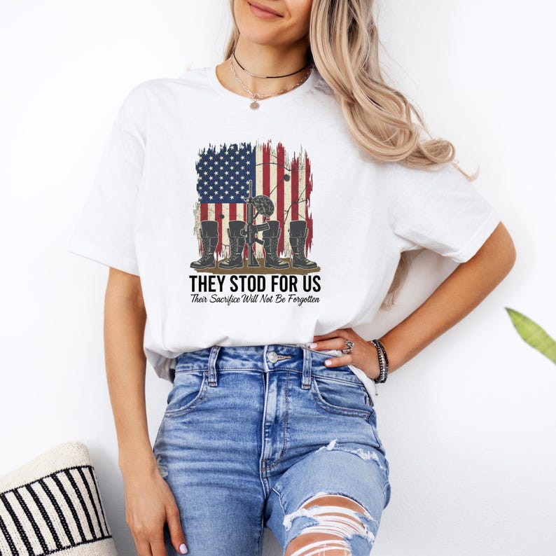 May include: White t-shirt featuring a patriotic design with an American flag and military boots. The text "THEY STOD FOR US" and "Their Sacrifice Will Not Be Forgotten" is printed below the graphic. A casual, everyday wear tee.