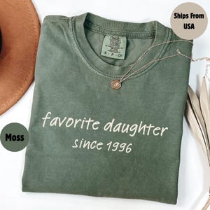 May include: A moss green t-shirt with the text "favorite daughter since 1996" in white. A silver necklace with a circular pendant rests on the shirt. The shirt is folded on a white surface.