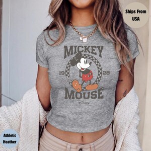 May include: Athletic heather gray t-shirt featuring a distressed graphic of Mickey Mouse. The design includes the words "Mickey Mouse" and the years "1928" and "19". Mickey is depicted in his classic outfit. The shirt is a crew neck.