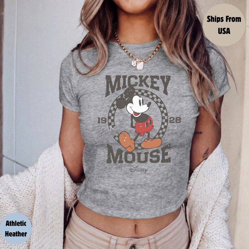 May include: Athletic heather gray t-shirt featuring a distressed graphic of Mickey Mouse. The design includes the words "Mickey Mouse" and the years "1928" and "19". Mickey is depicted in his classic outfit. The shirt is a crew neck.
