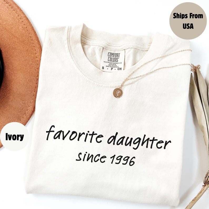 May include: Ivory-colored t-shirt with the text "favorite daughter since 1996" in black. A gold necklace with a pendant rests on the shirt. The shirt is folded, and a brown hat is partially visible.