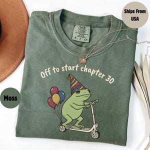 May include: Moss green t-shirt with the text "Off to start chapter 30". The graphic features a frog on a scooter with balloons. The shirt has a crew neck and short sleeves. A silver necklace is draped across the shirt.