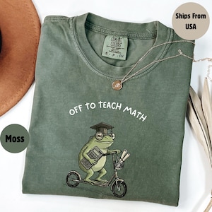 May include: A moss green t-shirt with the text "OFF TO TEACH MATH" and a cartoon frog wearing a graduation cap and glasses, riding a scooter while holding a calculator. A silver necklace is draped across the shirt.