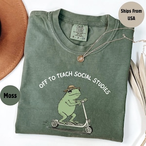 May include: Moss green t-shirt with the phrase "Off to Teach Social Studies" and a cartoon frog riding a scooter. The frog wears a hat. A gold necklace is draped across the shirt.