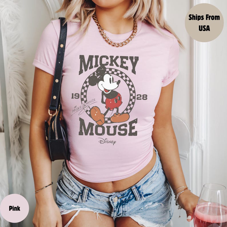 May include: Pink t-shirt featuring a distressed graphic of Mickey Mouse. The design includes the words "Mickey Mouse" and the year "1928" within a circular frame. The shirt is short-sleeved and has a crew neck.