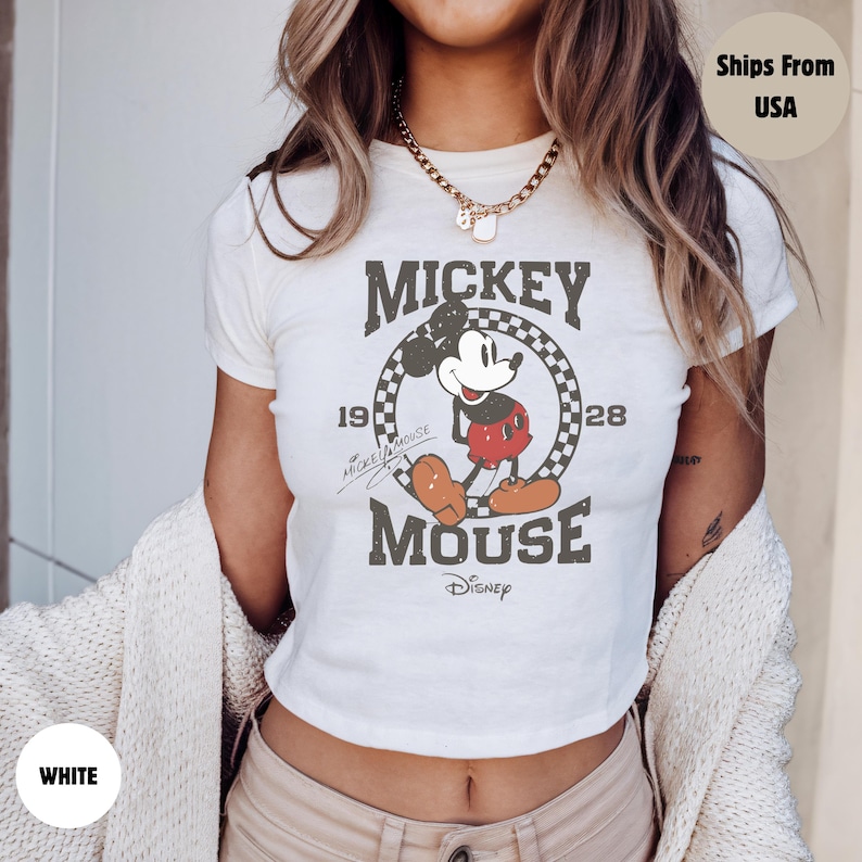 May include: White crop top featuring a distressed graphic of Mickey Mouse. The design includes the text "Mickey Mouse" and the years "1928" and "19". The Disney logo is also visible.