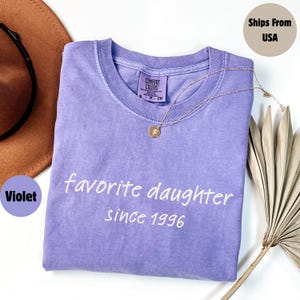 May include: A violet Comfort Colors t-shirt with the text "favorite daughter since 1996" in white. A gold necklace with a pendant rests on the shirt. A brown hat and dried plant stems are in the background.