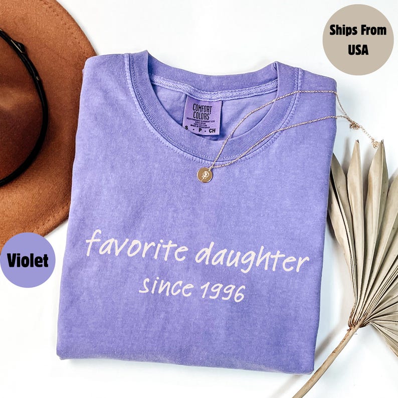 May include: A violet Comfort Colors t-shirt with the text "favorite daughter since 1996" in white. A gold necklace with a pendant rests on the shirt. A brown hat and dried plant stems are in the background.