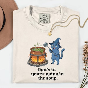 May include: Cream-colored t-shirt featuring a cartoon cat wearing a witch hat, holding a ladle over a cauldron of bubbling green soup with eyeballs. The text "that's it. you're going in the soup" is printed below the illustration. A gold necklace and a brown hat are also visible.