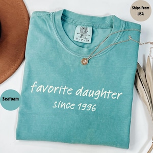 May include: Seafoam green t-shirt with the words "favorite daughter since 1996" in white script. A gold necklace with a pendant rests on the shirt. The shirt is folded and has a Comfort Colors tag.