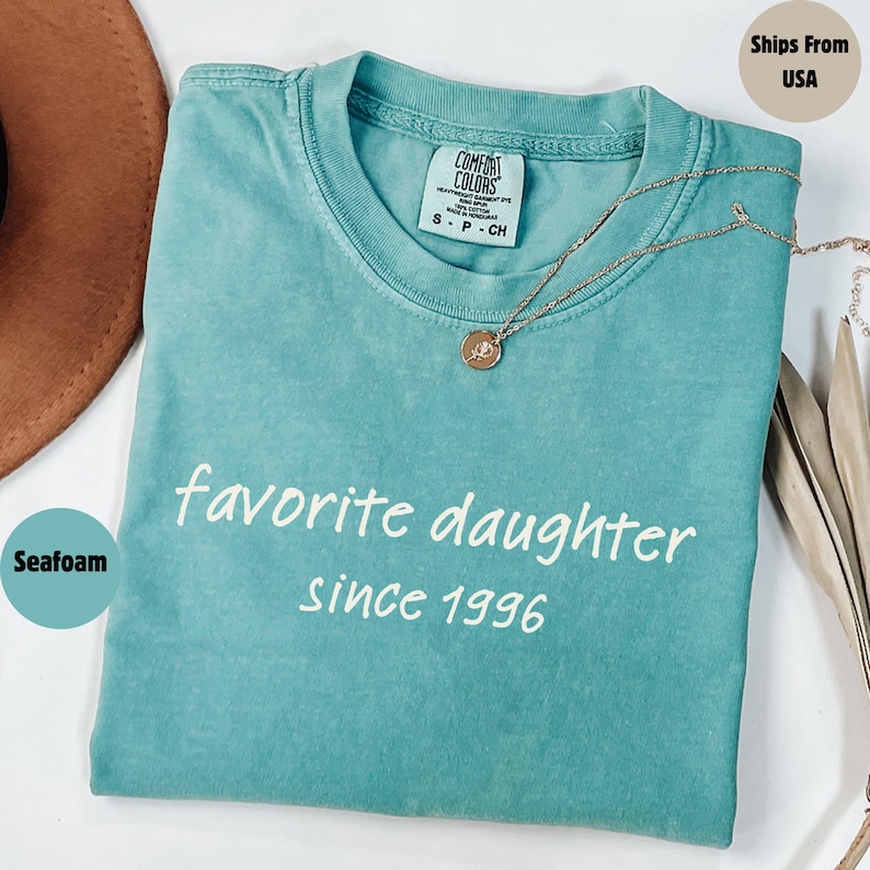 May include: Seafoam green t-shirt with the words "favorite daughter since 1996" in white script. A gold necklace with a pendant rests on the shirt. The shirt is folded and has a Comfort Colors tag.