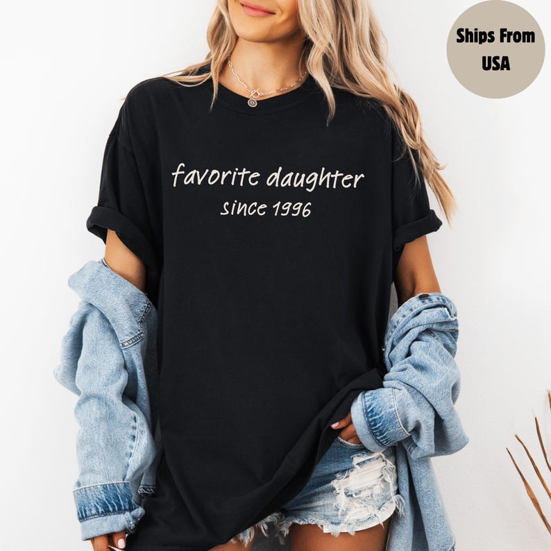 May include: Black t-shirt with the text "favorite daughter since 1996" in white script. The shirt is worn with a denim jacket and shorts. The model is wearing a necklace.