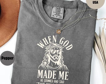May include: A gray Comfort Colors t-shirt with the text "WHEN GOD MADE ME HE GRIMMED AND SAID 'THIS WILL BE FUN'" in white. The graphic depicts a stylized Jesus wearing sunglasses. A silver necklace with a pendant rests on the shirt.