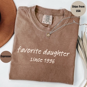 May include: Espresso-colored t-shirt with the text "favorite daughter since 1996" in white. The shirt has a crew neck and short sleeves. A silver necklace with a pendant is draped across the shirt.