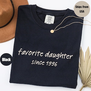 May include: Black t-shirt with the words "favorite daughter since 1996" in white script. A gold necklace with a small pendant rests on the shirt. A brown hat is visible in the upper left corner.