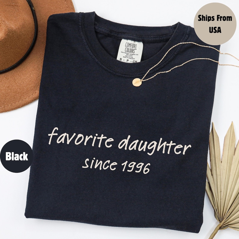 May include: Black t-shirt with the words "favorite daughter since 1996" in white script. A gold necklace with a small pendant rests on the shirt. A brown hat is visible in the upper left corner.