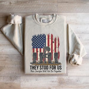 May include: Cream-colored sweatshirt featuring a distressed American flag design with a rifle, helmet, and boots. The text reads "THEY STOOD FOR US" and "Their Sacrifice Will Not Be Forgotten."