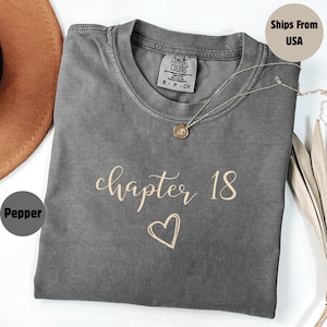 May include: A gray Comfort Colors t-shirt with the words "chapter 18" in a cursive font and a heart graphic. A silver necklace with a pendant is draped on the shirt. The shirt is a size small.