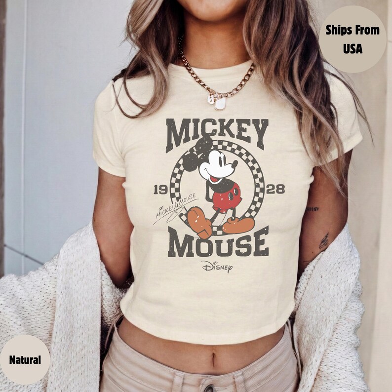 May include: Cream-colored t-shirt featuring a distressed graphic of Mickey Mouse. The design includes the words "Mickey Mouse" and the years "1928" and "19". The Disney logo is at the bottom. The shirt has short sleeves and a crew neck.