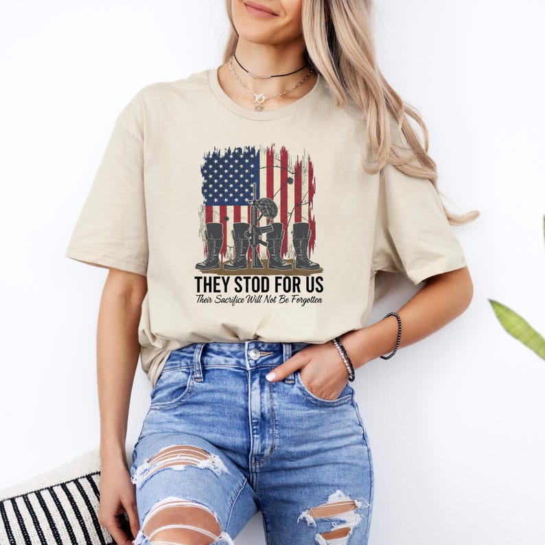 May include: Beige t-shirt featuring a graphic of the American flag with a distressed effect, a soldier's helmet, boots, and the text "THEY STOD FOR US". The text "Their Sacrifice Will Not Be Forgotten" is below the main text.