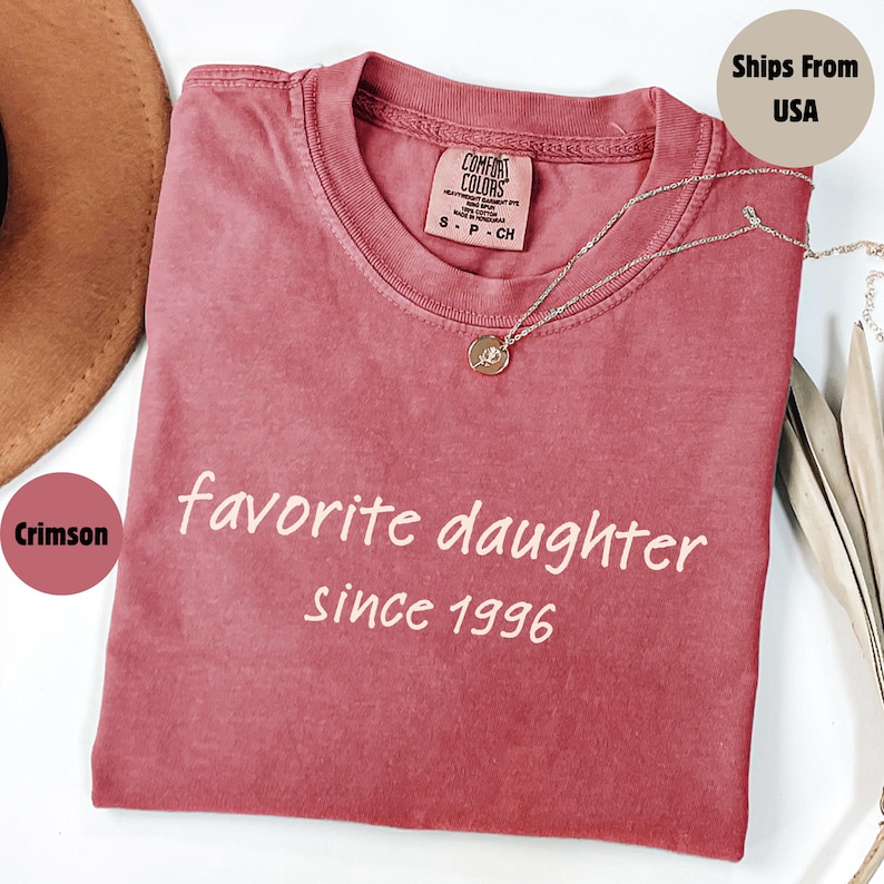 May include: Crimson-colored t-shirt with the text "favorite daughter since 1996" in white. A silver necklace with a pendant rests on the shirt. The shirt has a crew neck and a tag that reads "Comfort Colors".