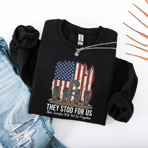 May include: Black sweatshirt featuring a distressed American flag design with a soldier's boots, rifle, and helmet. The text reads "THEY STOOD FOR US" and "Their Sacrifice Will Not Be Forgotten". A silver necklace is draped across the sweatshirt.