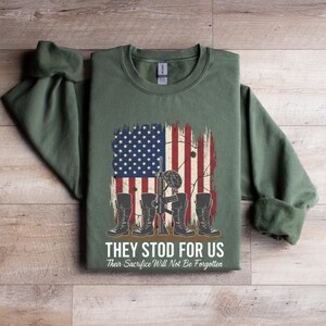 May include: Green sweatshirt with a distressed American flag graphic. The design includes military boots, a rifle, and the text "THEY STOD FOR US" and "Their Sacrifice Will Not Be Forgotten".