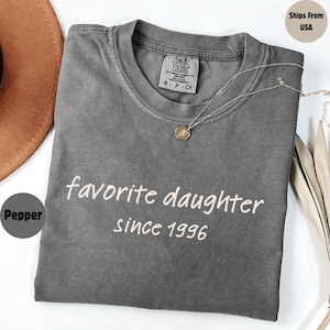May include: Pepper-colored Comfort Colors t-shirt with the text "favorite daughter since 1996" in white. A silver necklace with a pendant is draped on the shirt. The shirt is folded and displayed on a white surface.