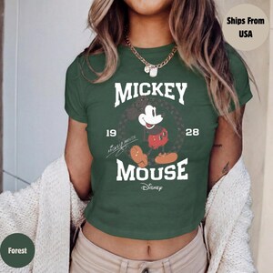 May include: Forest green crop top featuring a Mickey Mouse graphic. The design includes the words "Mickey Mouse" and the years "19" and "28". The Disney logo is at the bottom. The shirt has short sleeves and a crew neck.