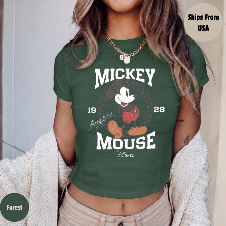 May include: Forest green crop top featuring a Mickey Mouse graphic. The design includes the words "Mickey Mouse" and the years "19" and "28". The Disney logo is at the bottom. The shirt has short sleeves and a crew neck.