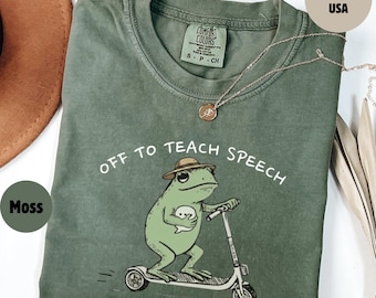 May include: A moss green t-shirt with the phrase "OFF TO TEACH SPEECH" and a cartoon frog riding a scooter. The frog wears a hat and holds a speech bubble. A gold necklace is draped on the shirt.