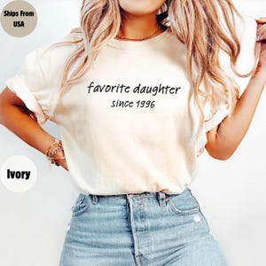 May include: Ivory-colored t-shirt with the text "favorite daughter since 1996" in black script. The shirt has short sleeves and is paired with blue denim jeans. The image shows a close-up of the shirt.
