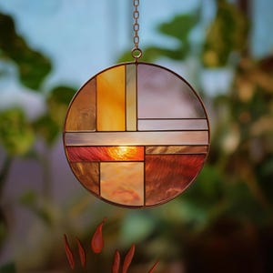 May include: A round stained glass suncatcher with a geometric design. The suncatcher features a variety of colors, including orange, yellow, red, and silver, and is suspended by a chain. The design is reminiscent of mid-century modern art.