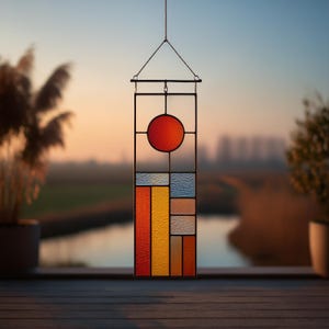 May include: A rectangular stained glass panel with a geometric design. The top features a red circle, and the lower section has vertical rectangles in shades of orange, yellow, blue, and beige. The panel is framed in black metal and hangs from a chain.