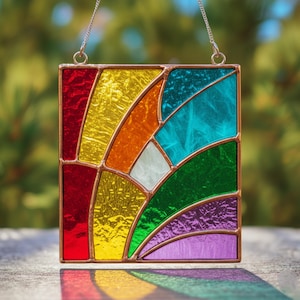 May include: A square stained glass panel with a rainbow design. The panel features sections of red, yellow, orange, white, turquoise, green, and purple glass, framed with copper. A silver chain is attached for hanging.