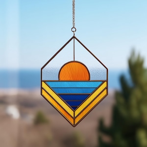May include: Stained glass suncatcher with a geometric design. The suncatcher features an orange sun, blue and yellow stripes, and a brown metal frame. It hangs from a chain and is displayed against a blurred background.