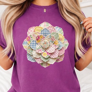May include: A berry-colored t-shirt featuring a large, colorful floral design. The flower is made of various patterned fabrics, including gingham and floral prints. The word "Berry" is printed in a script font at the bottom left of the shirt.