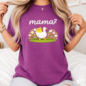 May include: A purple t-shirt featuring the text "mama&sup2;" above an illustration of a white duck with two yellow ducklings. The duck is walking on green grass surrounded by flowers.