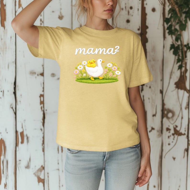 May include: Pale yellow t-shirt with the word "mama?" in white script. The shirt features a graphic of a white duck with two yellow ducklings, surrounded by flowers and grass.