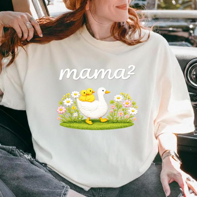 May include: Cream-coloured t-shirt with the white script text "mama&sup2;". The graphic below shows a white duck with two yellow ducklings, surrounded by flowers and grass. The t-shirt is a light colour.