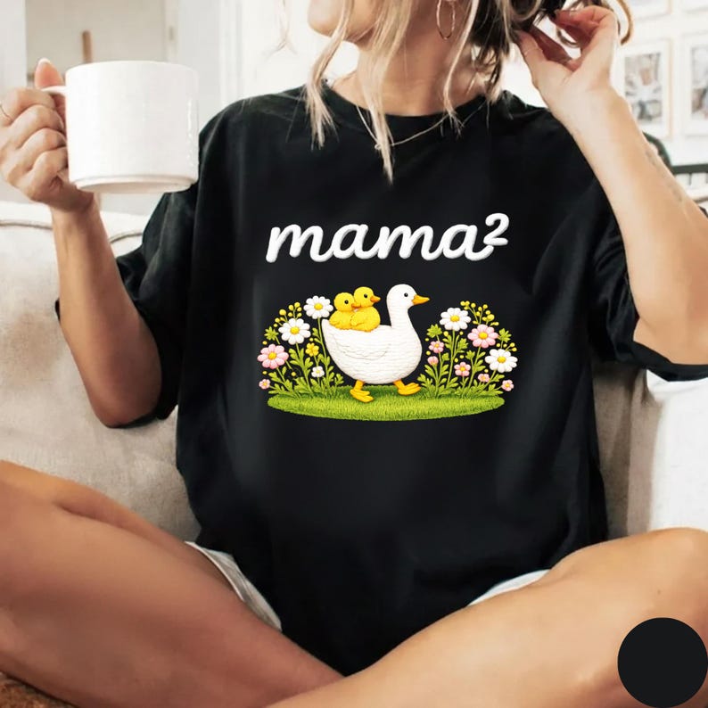 May include: Black t-shirt featuring the word "mama&sup2;" in white. The design includes a white duck with two yellow ducklings, set amidst flowers and grass. The t-shirt is a relaxed fit.