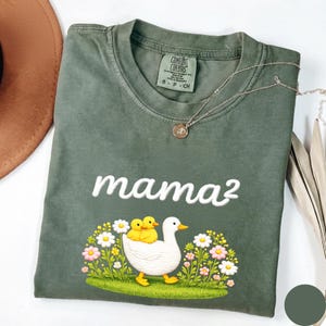 May include: A sage green Comfort Colors t-shirt with the embroidered word "mama&sup2;". The shirt displays a graphic of a white duck with two yellow ducklings, surrounded by flowers. A silver necklace with a pendant is draped across the shirt.