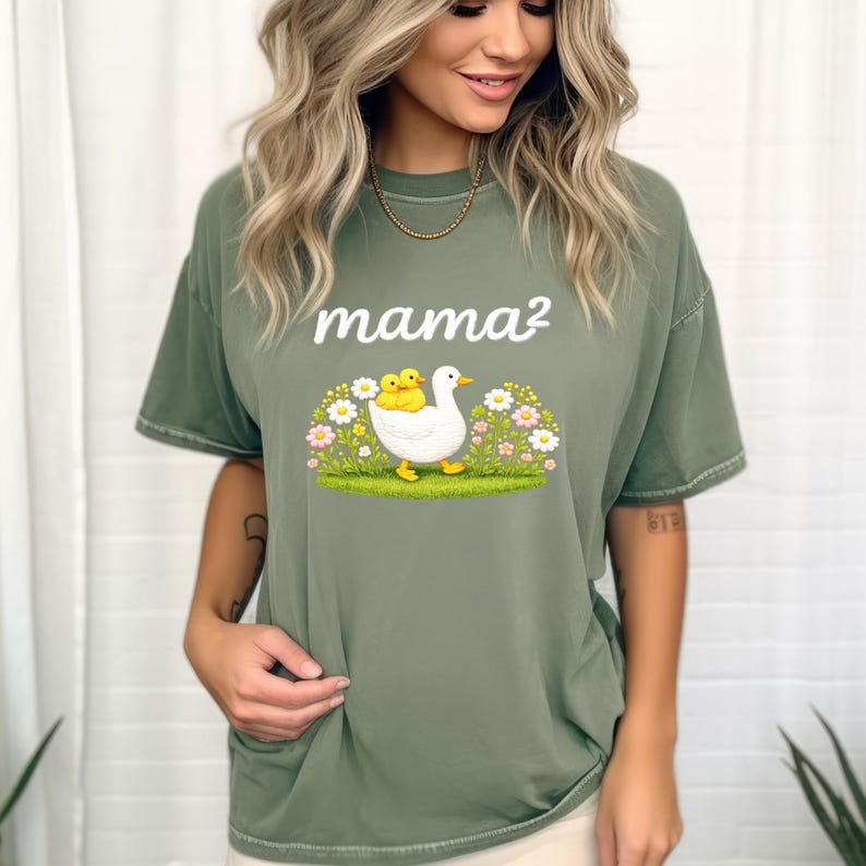 May include: Olive green t-shirt featuring the text "mama&sup2;" above a graphic of a white duck with two yellow ducklings. The duck is walking through a field of flowers and grass. The shirt has short sleeves.