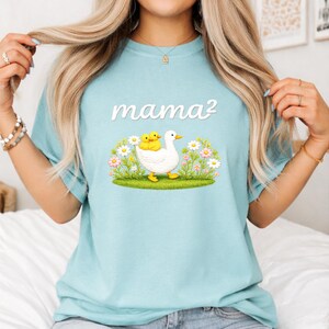May include: Pale blue t-shirt with the text "mama&sup2;" in white. The graphic shows a white duck with two yellow ducklings, set in a field of flowers and grass. The shirt is made of a soft, comfortable material.