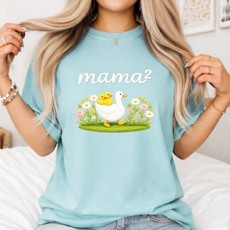 May include: Pale blue t-shirt with the text "mama&sup2;" in white. The graphic shows a white duck with two yellow ducklings, set in a field of flowers and grass. The shirt is made of a soft, comfortable material.