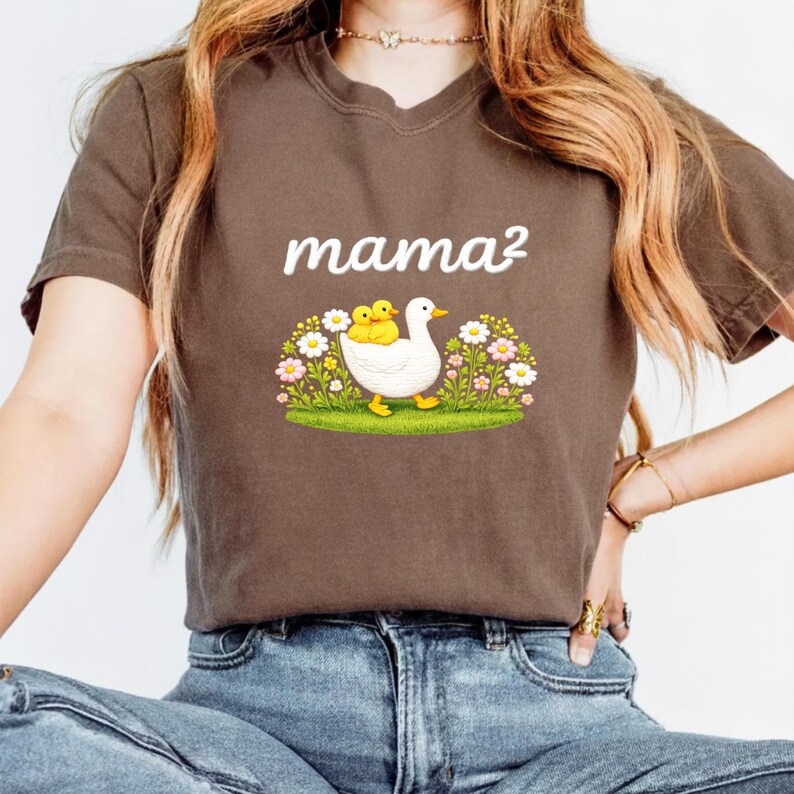 May include: A brown t-shirt with the text "mama&sup2;" in white script. The graphic shows a white duck with two yellow ducklings, set in a field of flowers and grass.