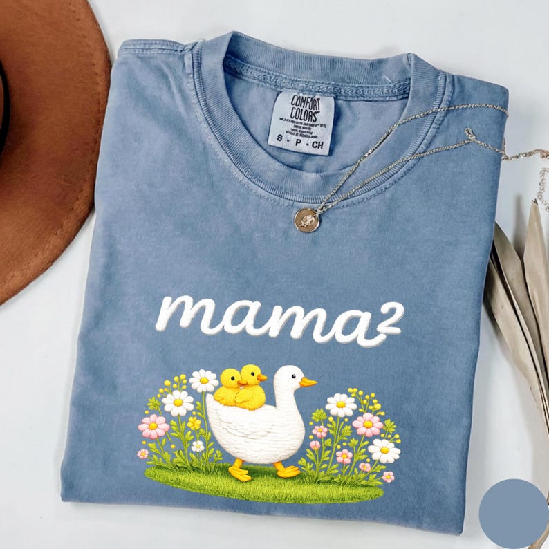 May include: A blue t-shirt with the embroidered text "mama&sup2;". The shirt displays a graphic of a white duck with two yellow ducklings, set amongst flowers and grass. A silver necklace with a pendant is also visible.